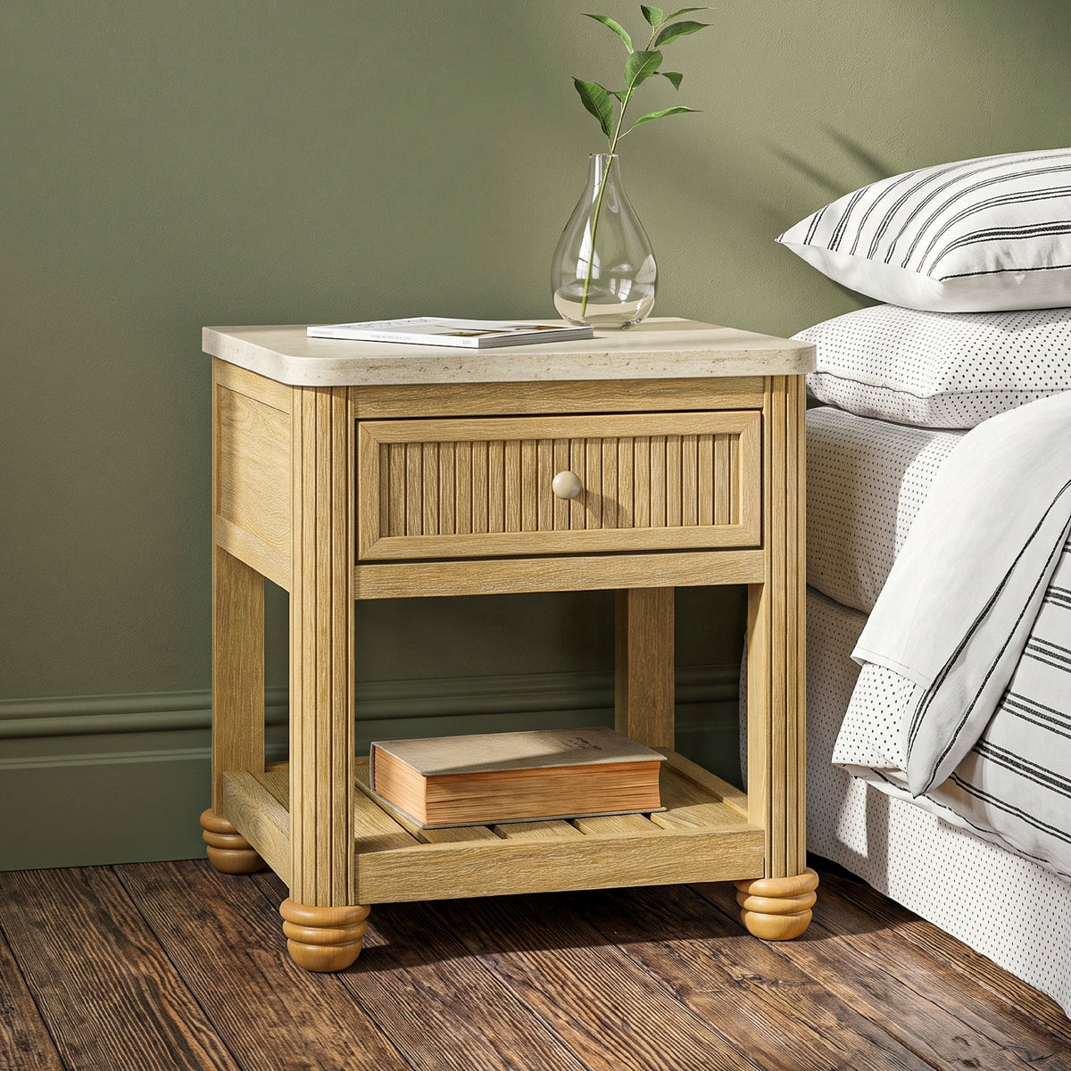 bedside table with drawer and open shelf in warm wood beside a bed against a green wall