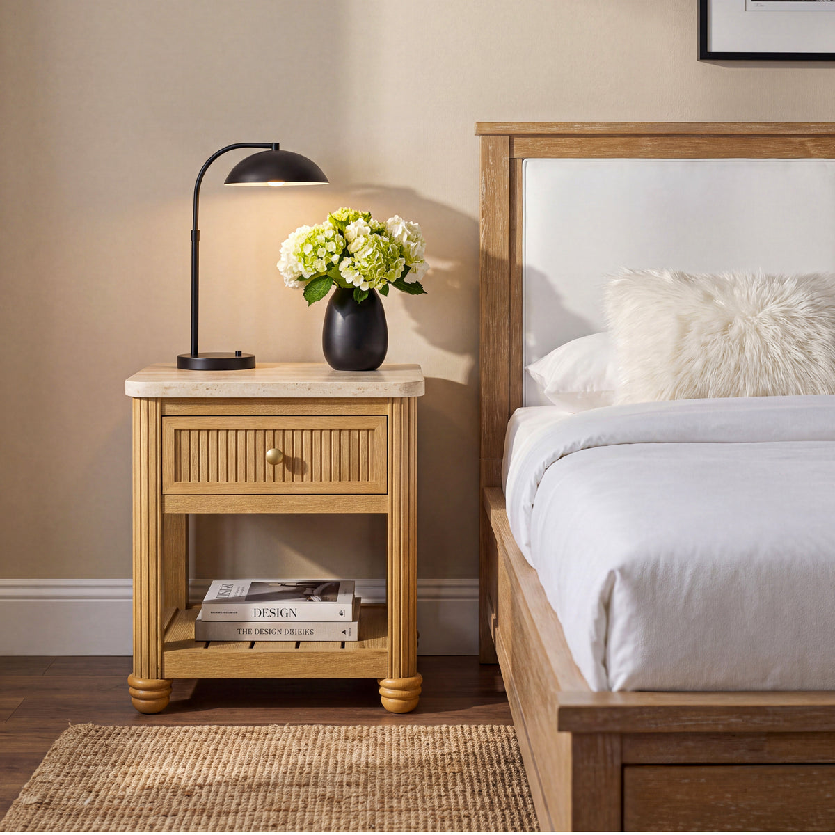 small nightstand with drawer and shelf styled with a black lamp and white flowers next to a bed